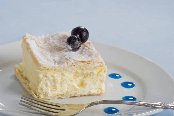 Quadrado de bolo de creme em um prato com mirtilos frescos em um fundo azul — Fotografia de Stock