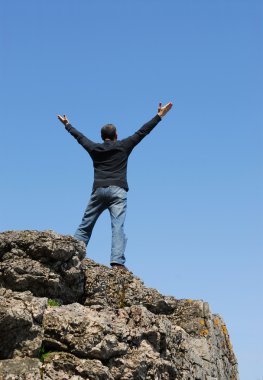 A man standing on a cliff with his arms raised to the blue sky clipart