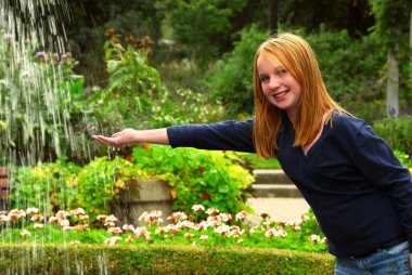 Young girl holding her hand under falling water in a garden clipart