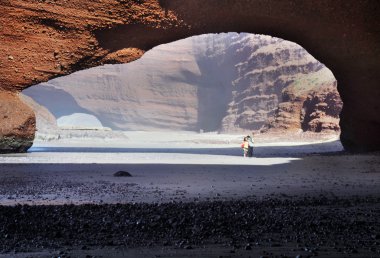 Young couple walking under arch rock formation near Atlantic in Morocco clipart