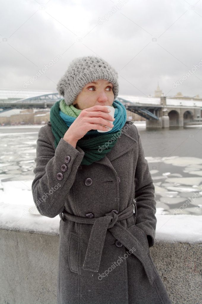 The young white frozen girl in a blue scarf, a gray cap and a coat drinks hot tea from a cup on Pushkinsky quay, Moscow, Russia in the winter