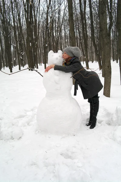 La muchacha abraza y besa al monigote de nieve grande en el parque en invierno —  Fotos de Stock