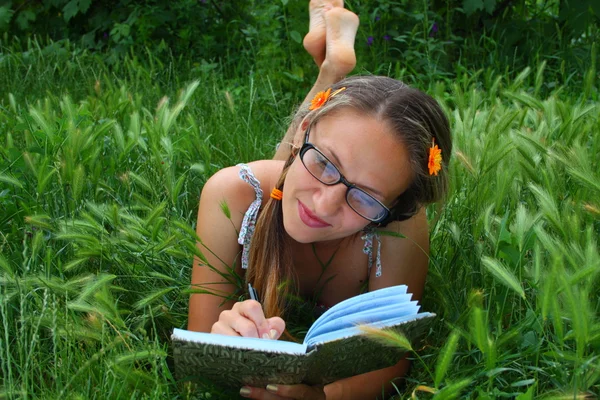 Vacker flicka i gräs leenden och skriver i en anteckningsbok — Stockfoto