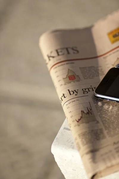 Close-up of financial newspaper and mobile phone, on a bench — Stock Photo, Image